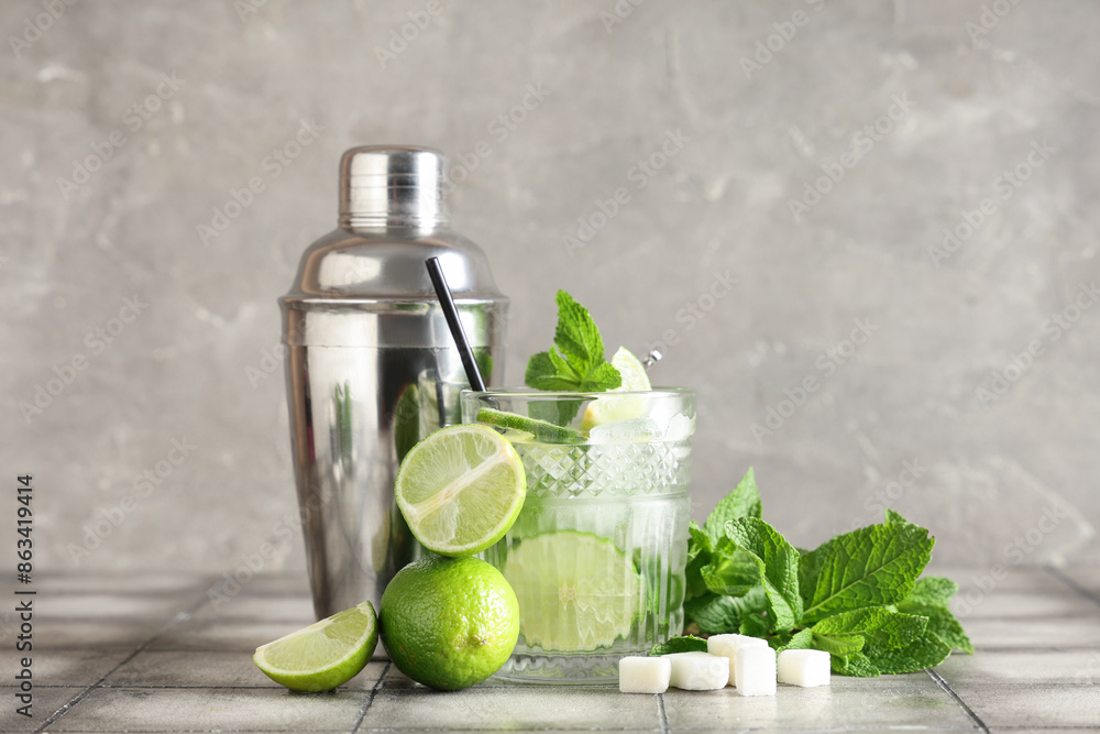 Glass of fresh Mojito cocktail with limes and shaker on grey tile table Glass of fresh Mojito cocktail with limes and shaker on grey tile table