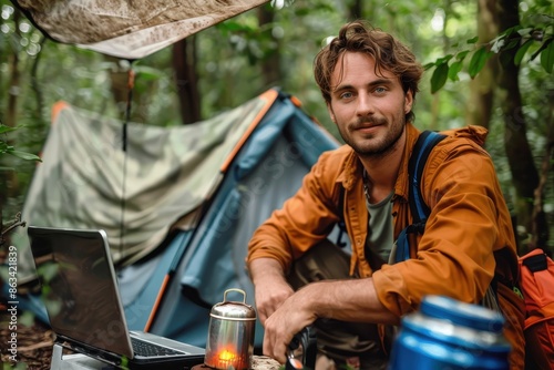 A man in an orange jacket is seated beside a tent in a lush forest, relaxing and using a laptop, with a pot nearby, suggesting a blend of work and wilderness adventure. photo