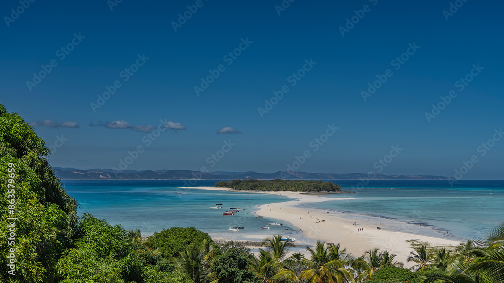 Obraz premium Beautiful postcard view. A sandbar meanders towards a tropical island with green vegetation. Tiny silhouettes of people on the beach. Boats in the aquamarine ocean. Palm leaves in the foreground.