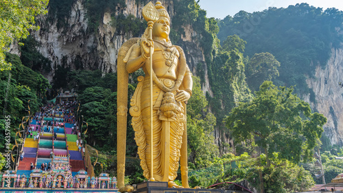 Many people climb the steps of the rainbow-colored staircase to the temple. Lush vegetation on the slopes of a sheer cliff. In the foreground- a giant golden statue of the god Lord Murugan. Batu caves photo