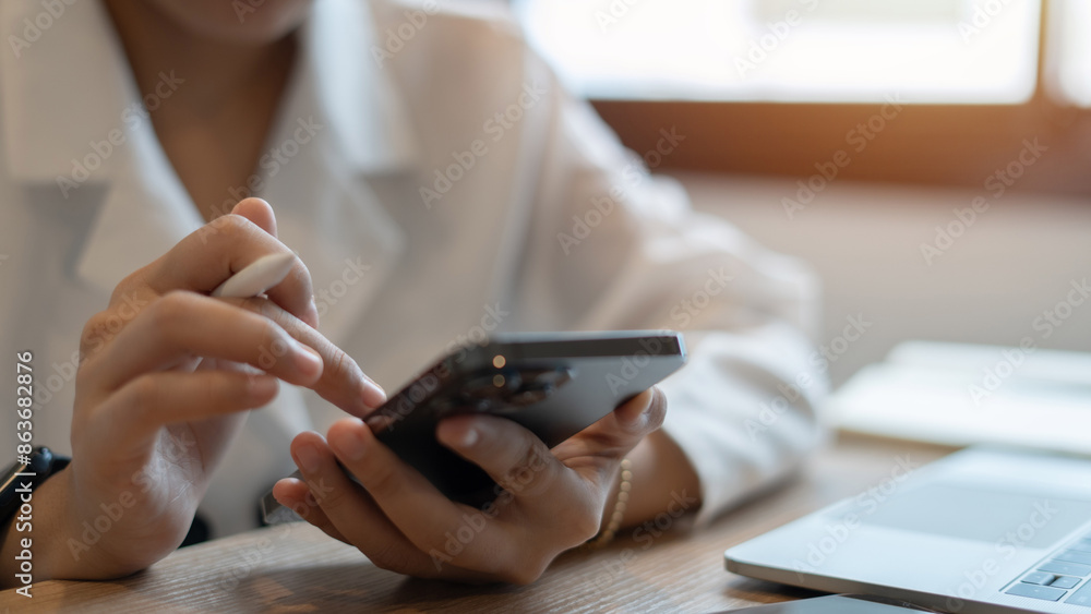 Fototapeta premium A woman is using her cell phone while sitting at a desk with a laptop. She is looking at the screen intently, possibly checking her email or browsing the internet. Concept of productivity and focus
