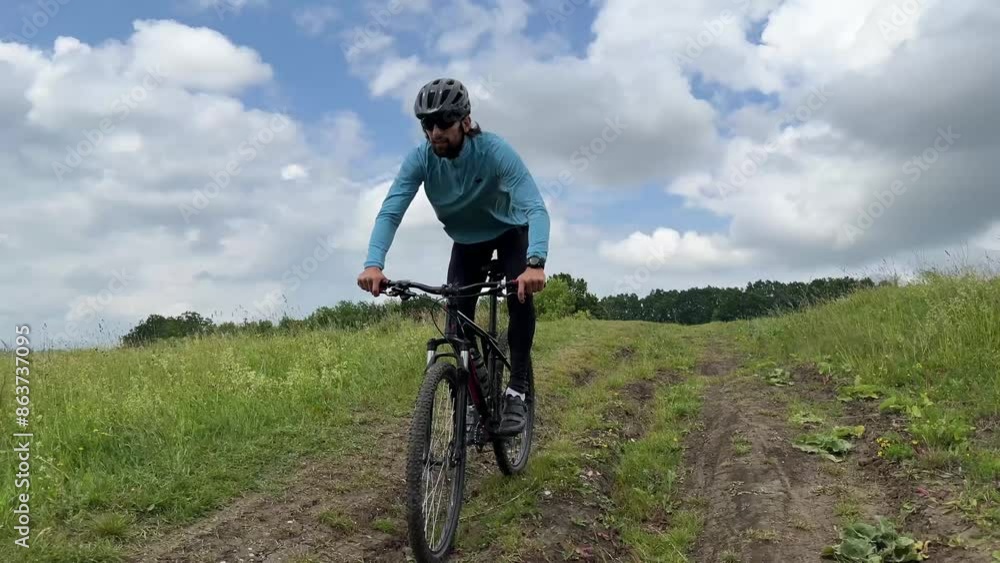 Mountain biker rides through lush countryside on a dirt trail, surrounded by green fields under a blue sky with fluffy clouds Mountain biker rides through lush countryside on a dirt trail, surrounded by green fields under a blue sky with fluffy clouds