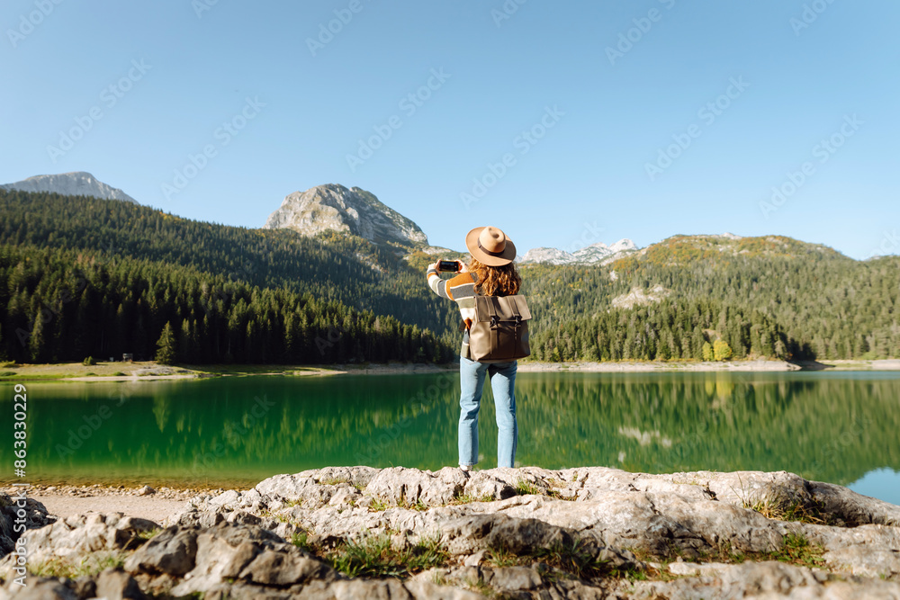 Obraz premium Young traveler is resting and taking pictures of amazing landscape with mountains and lake on smartphone during vacation. Tourism, nature, blogging concept.