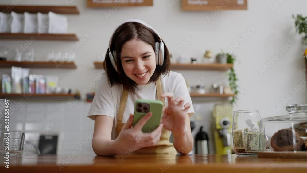 Happy brunette girl waiter in a yellow apron wearing white wireless headphones listens to music and watches a video during a break at work in a cafe Happy brunette girl waiter in a yellow apron wearing white wireless headphones listens to music and watches a video during a break at work in a cafe
