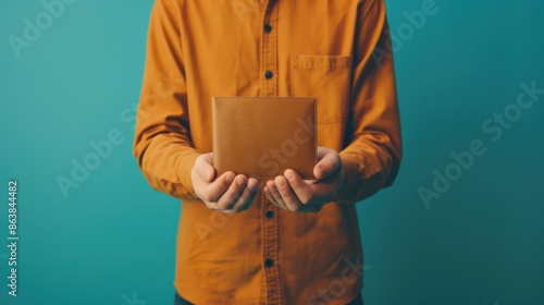 Person holding a brown leather wallet against a teal background, wearing an orange shirt. Minimalist style, close-up shot.