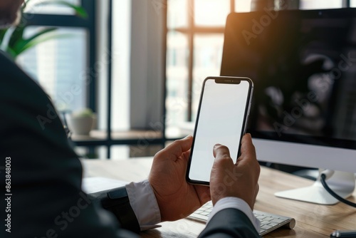 Mockup of a man's hands holding smart phone with blank white screen while sitting at the wooden table in modern office
