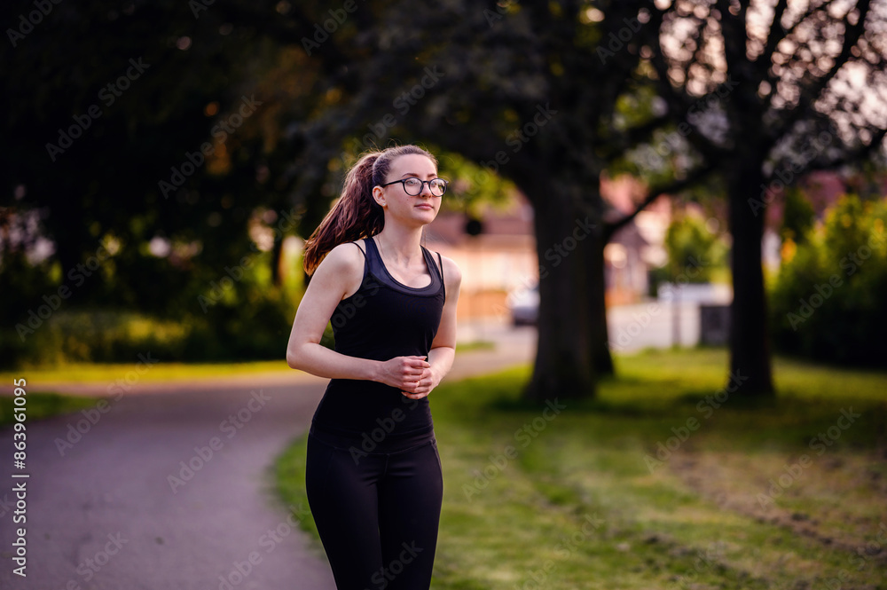 A young woman in black sportswear and glasses jogging on a park path at dusk, showcasing determination and a healthy lifestyle. A young woman in black sportswear and glasses jogging on a park path at dusk, showcasing determination and a healthy lifestyle.