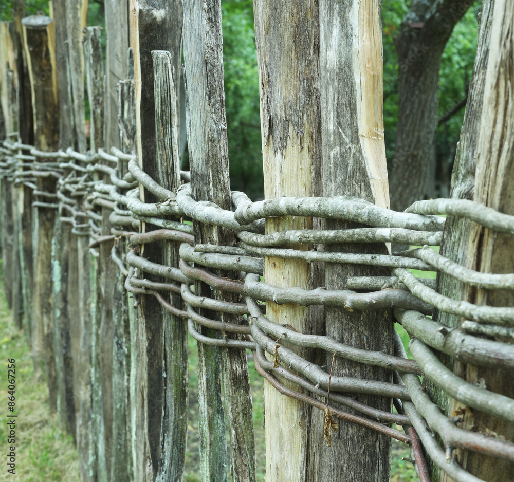 Fototapeta premium Wooden fence assembled of beams or logs old patched up woven wicker with bits of wood. Natural building concept. Green background nature