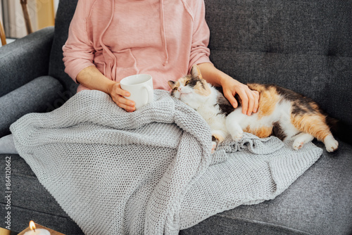 Photography No face woman in plaid drinking hot tea, petting a relaxed cat on the sofa at home