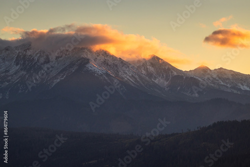 A beautiful autumn landscape of the Tatra Mountains seen from Polish Podhale during the golden hour. The sky is painted in warm hues, with the mountains bathed in golden light.