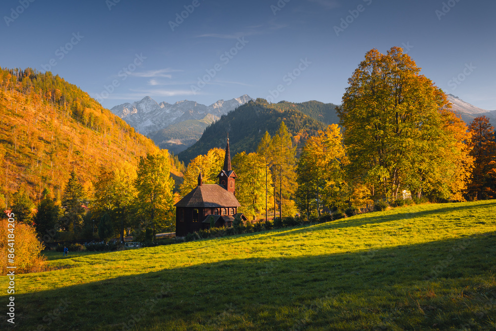 Obraz premium Autumn view of the Tatras in Slovakia: golden, red, and orange leaves on trees surround majestic mountains with snow-capped peaks. Clear, blue sky contrasts with the warm colors of the forest. 