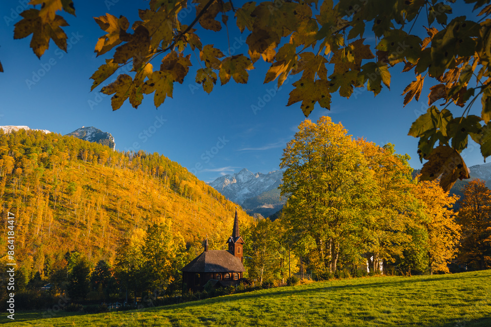 Autumn view of the Tatras in Slovakia: golden, red, and orange leaves on trees surround majestic mountains with snow-capped peaks. Clear, blue sky contrasts with the warm colors of the forest. Autumn view of the Tatras in Slovakia: golden, red, and orange leaves on trees surround majestic mountains with snow-capped peaks. Clear, blue sky contrasts with the warm colors of the forest.