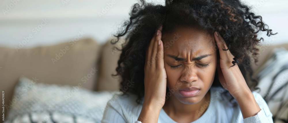 Young African American woman sitting on sofa, exhausted and holding her head in pain Young African American woman sitting on sofa, exhausted and holding her head in pain