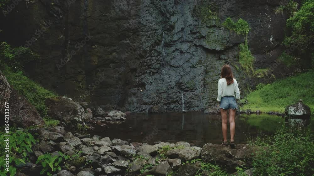 A lone woman stands on a rocky shore, admiring a cascading waterfall in the lush tropical landscape of French Polynesia. The water flows into a tranquil pool surrounded by verdant vegetation. A lone woman stands on a rocky shore, admiring a cascading waterfall in the lush tropical landscape of French Polynesia. The water flows into a tranquil pool surrounded by verdant vegetation.