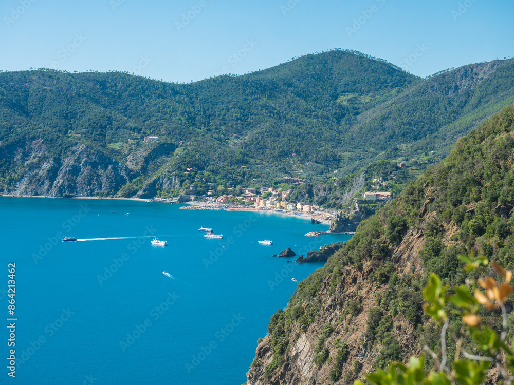 Fototapeta premium Picturesque distant view of Monterosso al Mare with beach, ships and boats seen from Azure Trail to Vernazza with green cliffs, rock, clear blue sky and sea. National park Cinque Terre, Liguria, Italy