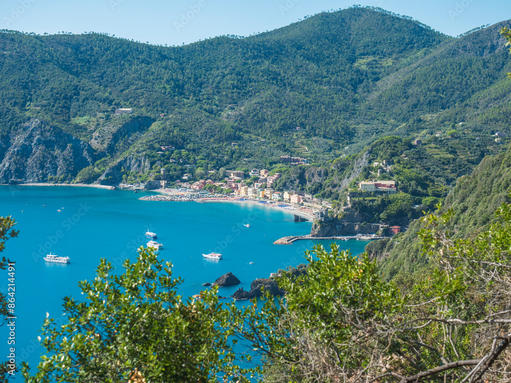 Fototapeta premium Picturesque distant view of Monterosso al Mare with beach, ships and boats seen from Azure Trail to Vernazza with green cliffs, rock, clear blue sky and sea. National park Cinque Terre, Liguria, Italy