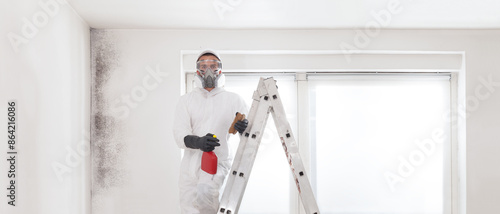 Professional mold removal service: Worker on ladder wearing protective suit, gloves, glasses and respirator mask. Holding spray bottle product for Cleaning moldy wall before room painting work.