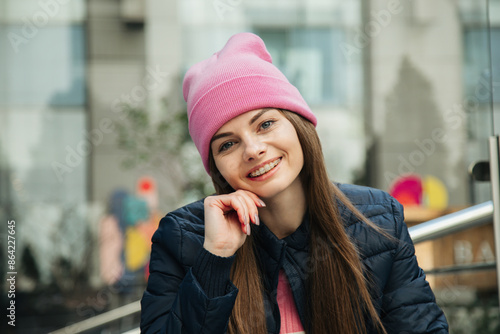 Smiling young woman with braces in a pink hat and blue jacket sitting on the street.