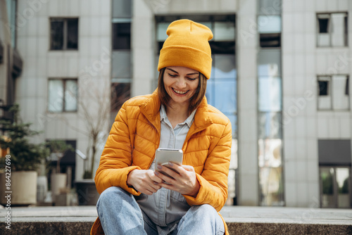Smiling woman in yellow jacket sitting on the street and holds the phone in her hands.