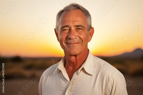 Portrait of a grinning man in his 60s wearing a simple cotton shirt while standing against vibrant sunset horizon
