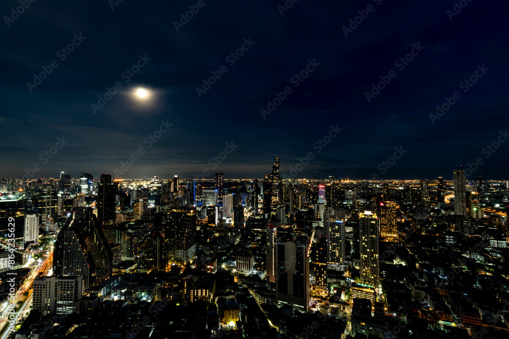 Obraz premium A breathtaking night view of Bangkok's skyline illuminated by city lights and a full moon. The cityscape showcases the blend of modern skyscrapers and vibrant urban life, reflecting the city's dynamic