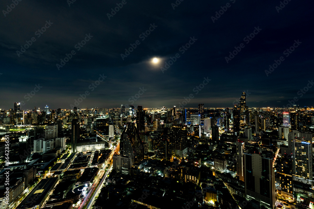 Obraz premium A breathtaking night view of Bangkok's skyline illuminated by city lights and a full moon. The cityscape showcases the blend of modern skyscrapers and vibrant urban life, reflecting the city's dynamic