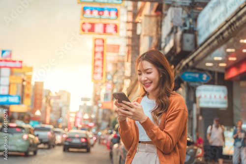 Happy young Asian tourist woman using smartphone on street, Female traveller in Yaowarat Chinatown street food market during sunset in Bangkok, Thailand. photo