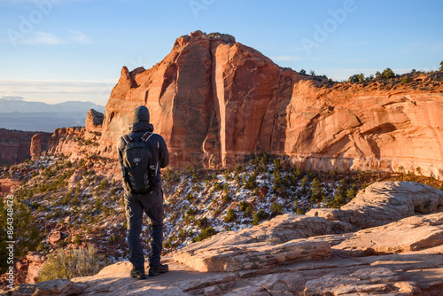 A male hiker stands at the edge of a cliff in Utah's Canyonlands National Park, admiring the breathtaking view of the snow-dusted landscape and rugged red rock formations at sunrise - USA