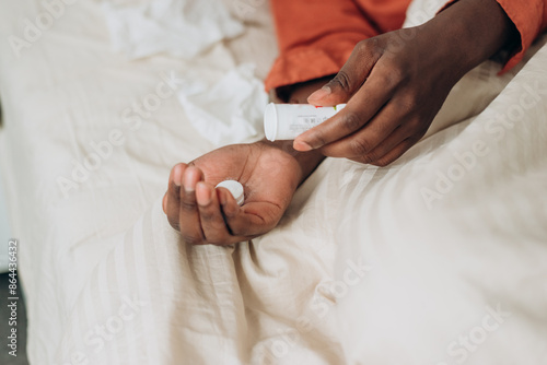 Close-up of African American man taking medication while lying in bed, depicting moment of self-care and health management. Male poured antipyretic pills into his hand. photo
