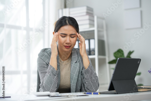 Businesswoman suffering from headache at work, sitting at desk with laptop, experiencing stress and fatigue in office environment