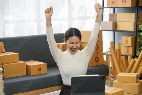 Excited woman celebrating success in online business with raised arms, surrounded by packages, looking at a laptop. photo