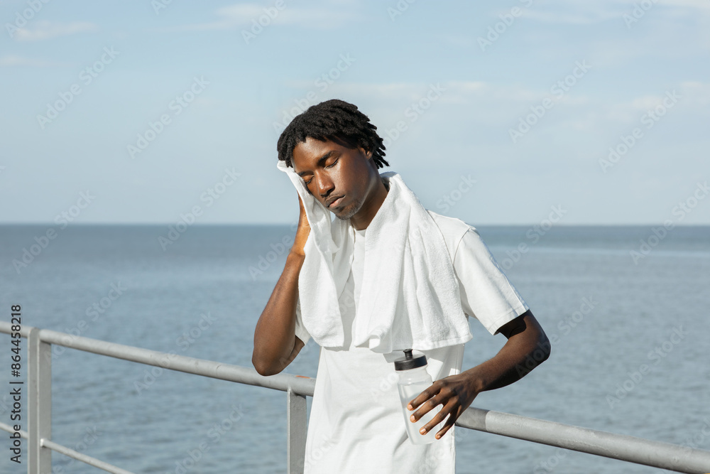 African American young man, likely an athlete, takes a break on a railing by the ocean. He is wearing a white t-shirt and towel and holding a water bottle. His face looks exhausted African American young man, likely an athlete, takes a break on a railing by the ocean. He is wearing a white t-shirt and towel and holding a water bottle. His face looks exhausted