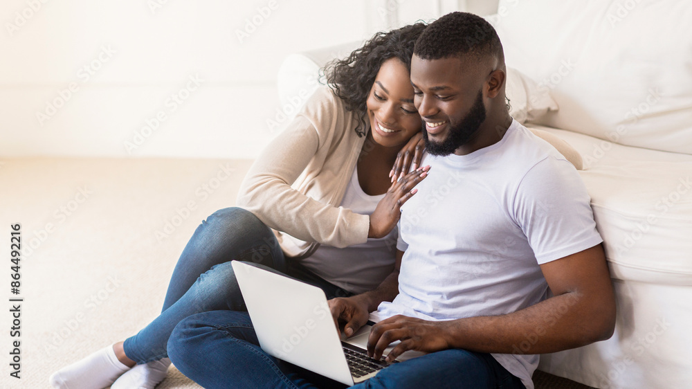A smiling couple sits on the floor of their living room, using a laptop together. The woman is sitting to the mans left and has her arm around his shoulder. A smiling couple sits on the floor of their living room, using a laptop together. The woman is sitting to the mans left and has her arm around his shoulder.