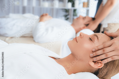 Caucasian couple enjoying relaxing anti-stress head massage and pampering facial beauty skin recreation leisure in dayspa modern light ambient at luxury resort or hotel spa salon. Quiescent