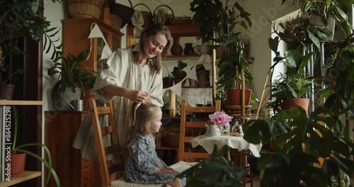 Wallpaper Mural Young loving mother tying a hair bow for her adorable little daughter in grey dress sitting on a wooden chair among decorative plants in the home garden. Wide slow motion shot. Torontodigital.ca