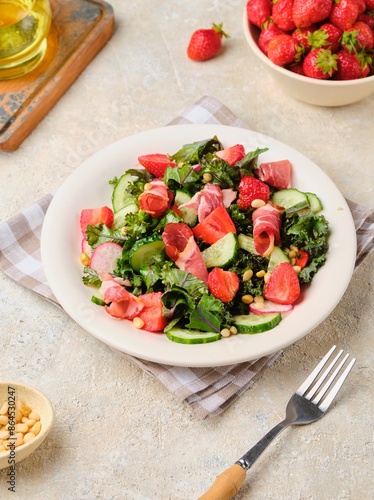 A healthy salad with cabbage kale, fresh cucumbers, radish, strawberries and smoked meat on a beige ceramic plate on a light concrete background.