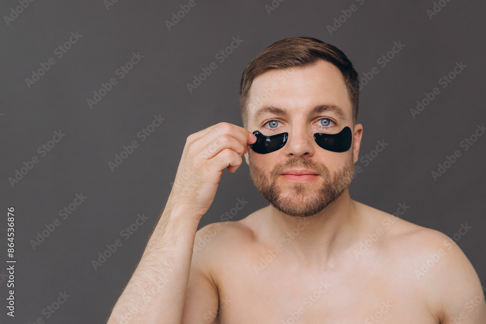 Close-up portrait of handsome bearded man applying black patches under eyes. Attractive guy caring about his appearance on grey background Close-up portrait of handsome bearded man applying black patches under eyes. Attractive guy caring about his appearance on grey background