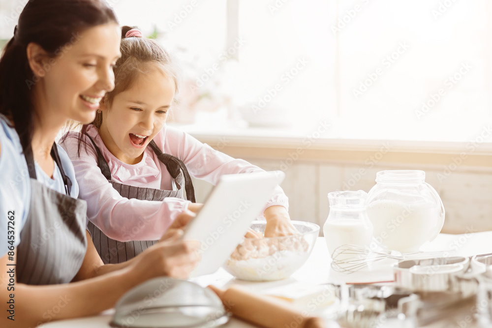 A mother and daughter are baking together in the kitchen. They are looking at a tablet, presumably for a recipe. There are various baking ingredients and tools on the table, copy space A mother and daughter are baking together in the kitchen. They are looking at a tablet, presumably for a recipe. There are various baking ingredients and tools on the table, copy space