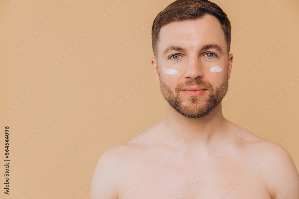 Obraz premium Attractive bearded man with healthy skin, applies cream moisturizer for anti wrinkle or anti aging, cares of body, poses naked or shirtless, isolated over beige background with free space.