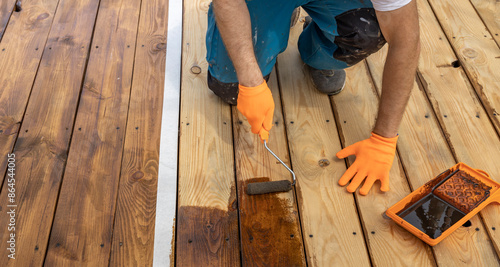 Wallpaper Mural A person wearing orange gloves is applying a wood stain to a deck with a roller. A tray of wood stain is nearby. Torontodigital.ca