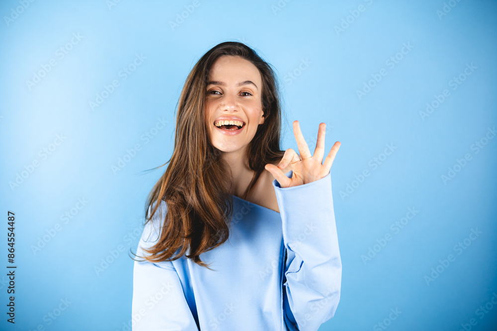 Happy young charming woman showing ok sign with fingers, trying to say that everything is super, joyfully looking at camera, isolated on blue background. Happy young charming woman showing ok sign with fingers, trying to say that everything is super, joyfully looking at camera, isolated on blue background.