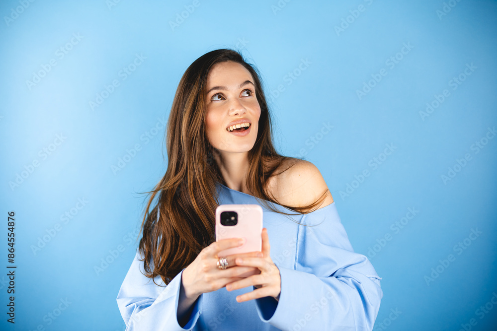 Smiling beautiful young woman in blue pullover isolated on blue background. Girl flirting while chatting on mobile phone, dreaming, look at side. Woman think about answer, dreaming. Smiling beautiful young woman in blue pullover isolated on blue background. Girl flirting while chatting on mobile phone, dreaming, look at side. Woman think about answer, dreaming.