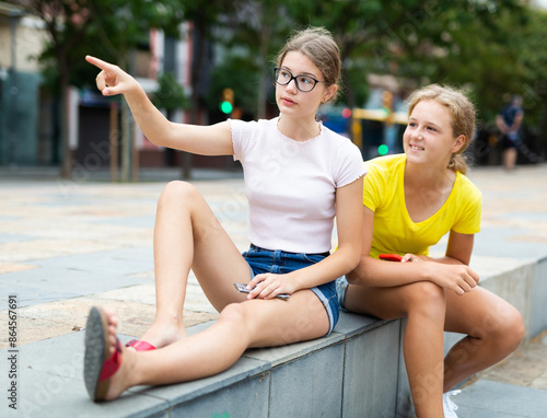 Two friends are sitting on street parapet. One of them points with her hand to something