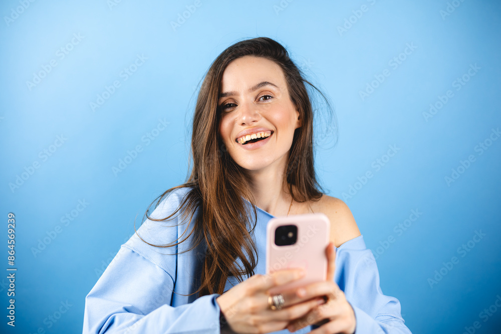 Attractive cheerful amazed girl using device gadget app, sms, post, share social media news isolated blue background. Positive woman holding gadget, texting, messaging. Girl wear blue pullover. Attractive cheerful amazed girl using device gadget app, sms, post, share social media news isolated blue background. Positive woman holding gadget, texting, messaging. Girl wear blue pullover.