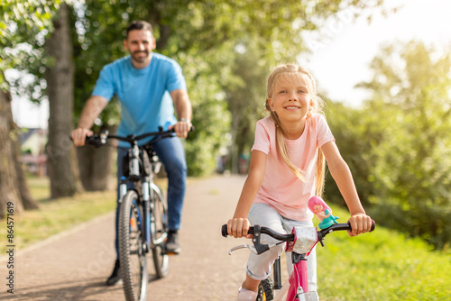 A young girl is smiling as she rides a pink bicycle on a paved path in a park. Her father is behind her, also riding a bicycle. Both are enjoying a sunny day outdoors. © Prostock-studio