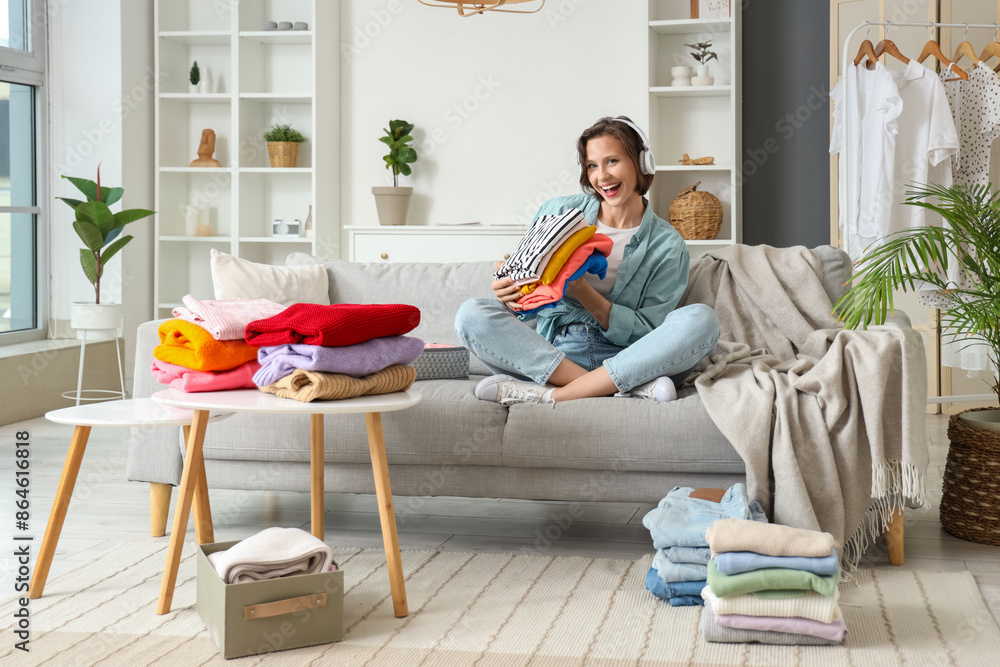 Young woman in headphones folding different clothes and sitting on sofa at home Young woman in headphones folding different clothes and sitting on sofa at home