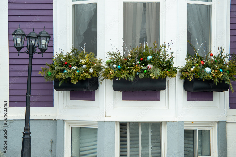 Fototapeta premium Three black window boxes filled with Christmas decor hung on the outside of a purple wooden house with white trim. The wood pots are filled with ornaments, plastic balls, and green spruce garlands. 