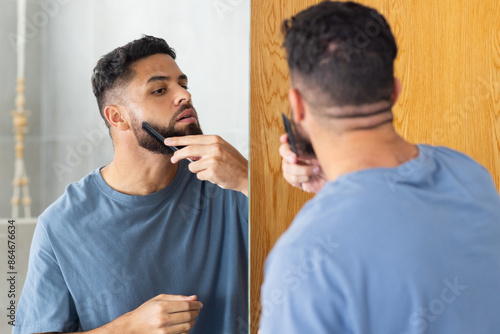 Grooming beard, man using comb and mirror for precise styling at home