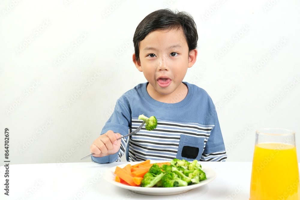 A young boy with a playful expression holds a fork with broccoli, sitting in front of a healthy meal and orange juice. This image is perfect for promoting healthy eating, children's nutrition, and bal
