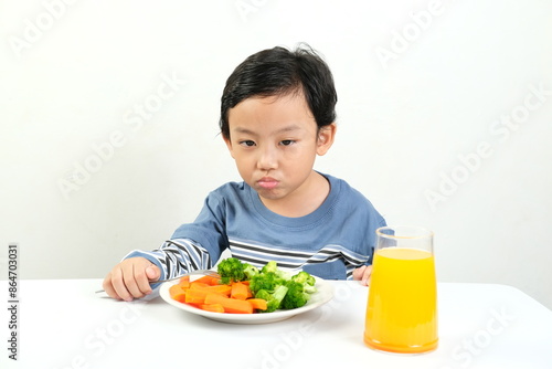 Fotografía A young boy with a pouty expression stares at a plate of vegetables and a glass of orange juice, showing clear reluctance